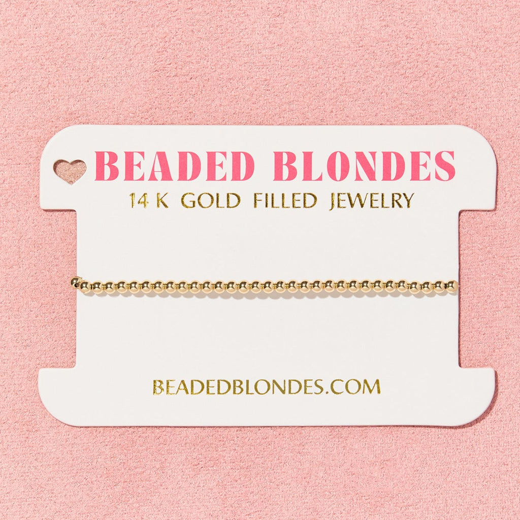Gold bracelet on a 'Beaded Blondes' branded card against a pink background