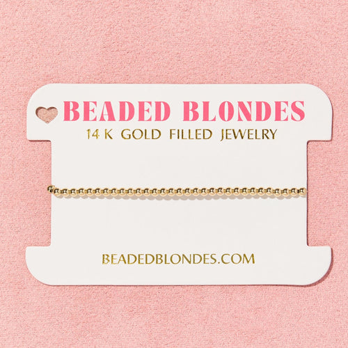 Gold bracelet on a 'Beaded Blondes' branded card against a pink background