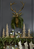 Decorative mantel with gold deer head, greenery, and wooden trees against a dark wall.