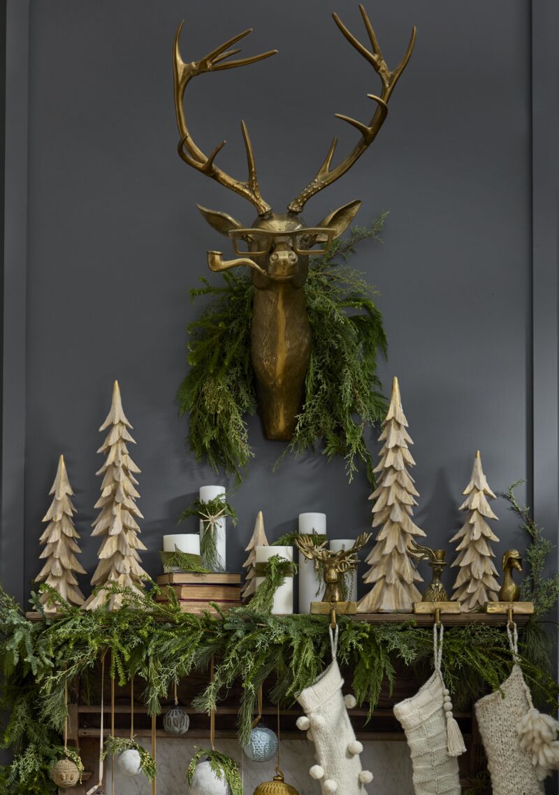 Decorative mantel with gold deer head, greenery, and wooden trees against a dark wall.