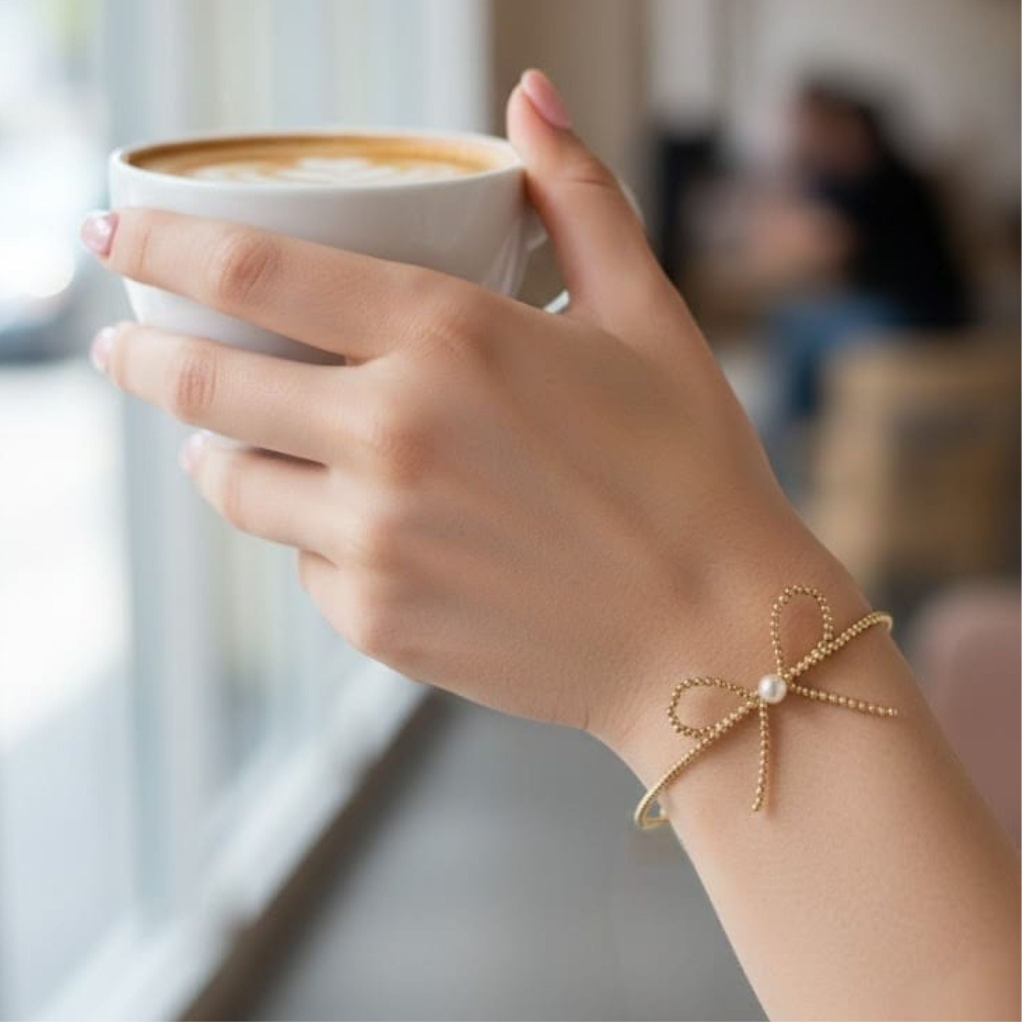 Hand holding a cup of coffee with a gold bracelet featuring a bow design.