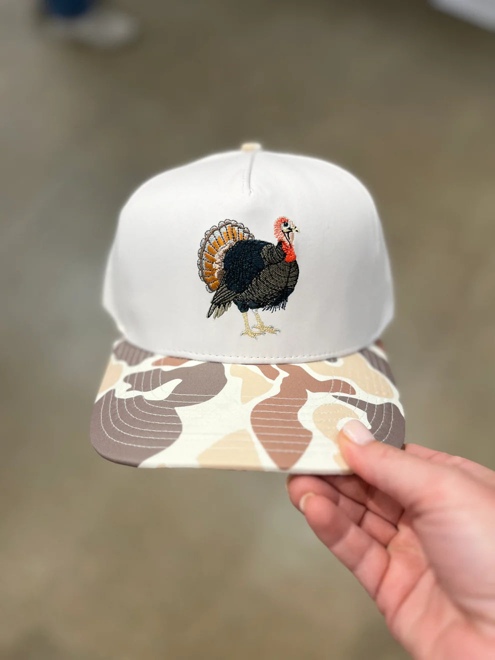 White cap with a turkey design on the front, held by a hand against a neutral background.
