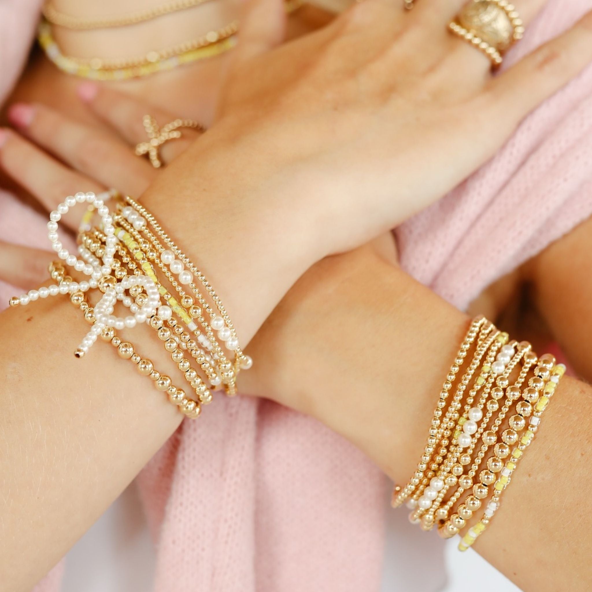 Close-up of hands wearing multiple gold bracelets on a light pink background