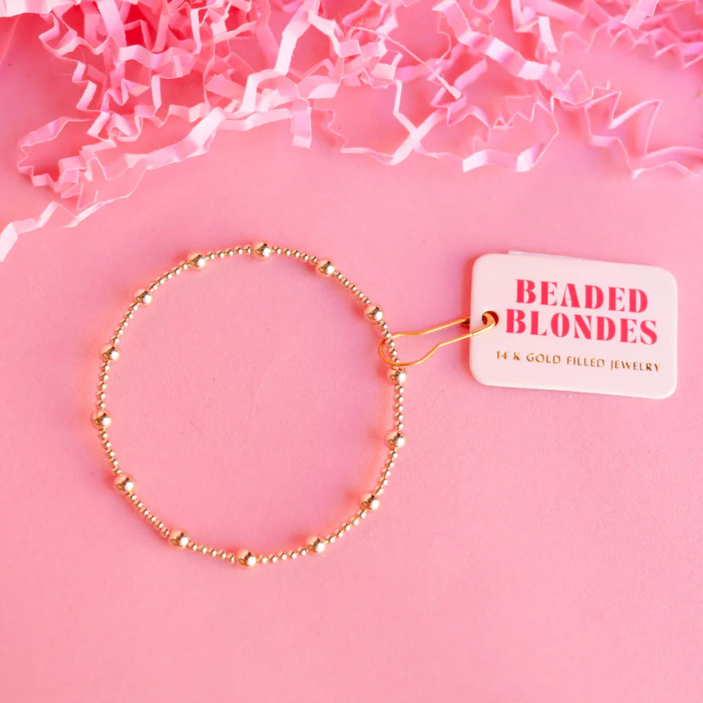 Gold beaded necklace with a 'Beaded Blondes' tag on a pink background