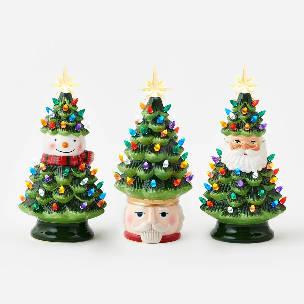 Three small ceramic Christmas tree figurines on a light gray background: one with a snowman head, one with a Santa Claus face in the middle, and one with a Santa Claus face on the right. The trees are green with multicolored lights and gold star toppers.