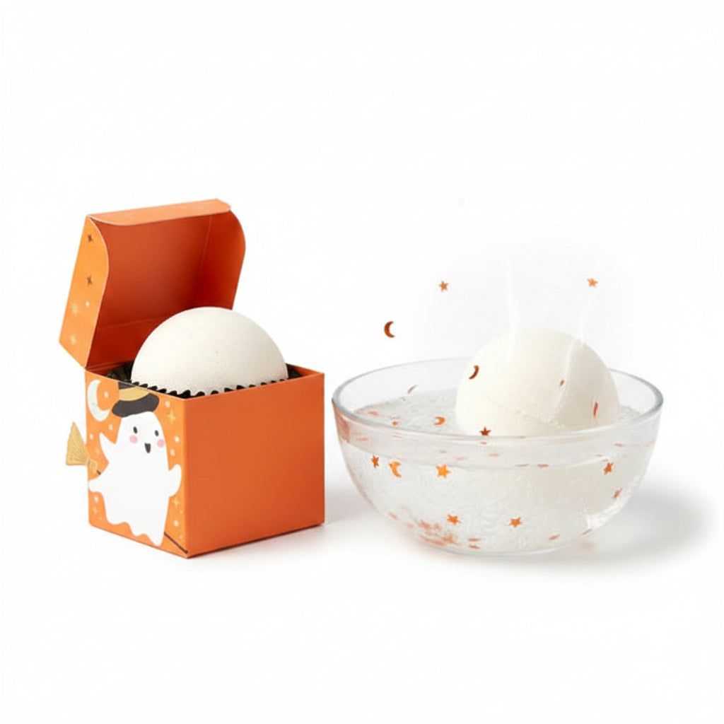 A Halloween-themed bath bomb set featuring a round off-white bath bomb next to its orange packaging box decorated with a cartoon ghost, witch hat, and broom.