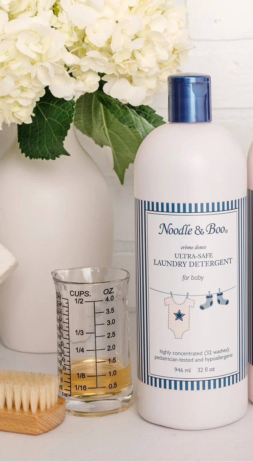 Two bottles of Noodle & Boo baby products on a surface with flowers and a measuring cup in the background.