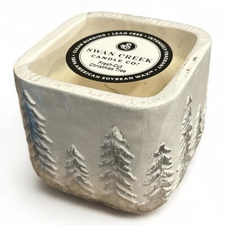 A square ceramic candle jar with a textured, rustic finish, featuring embossed pine tree designs on the sides. The top is open to reveal a cream-colored wax pool and a circular black and white label that reads 'Shan Creek Candle Co.