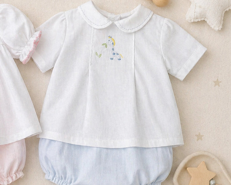Two baby girls' short-sleeve tops with matching bloomers laid flat on a soft beige surface. The left outfit is white with pale pink trim and a small pastel floral embroidery; the right outfit is white with pale blue trim and a small pastel floral embroidery.