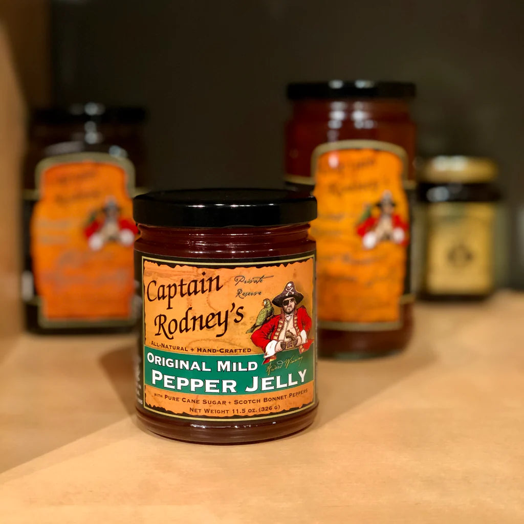 Jar of Captain Rodney's Original Mild Pepper Jelly on a wooden surface with other jars in the background.