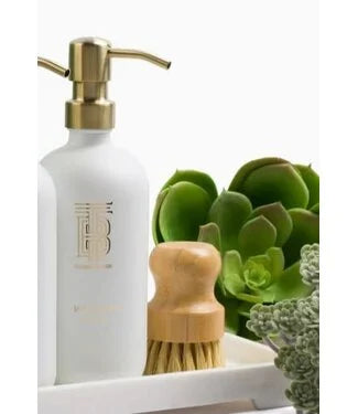 White soap dispenser with gold pump, wooden brush, and succulents on a white background